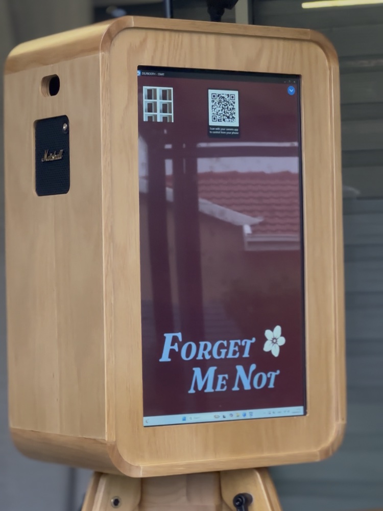 The Forget Me Not booth: a wooden box camera on a timber tripod, its screen
                    showing the Forget Me Not welcome.
