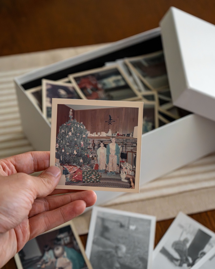 A hand holding an old photograph above an open box of family photographs.